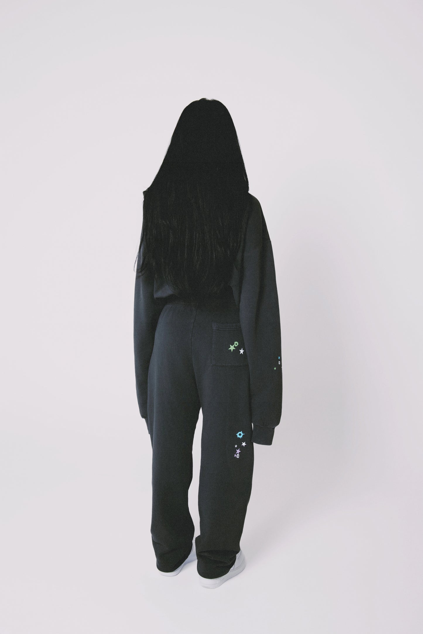 Back view of a model wearing black 'SERIAL' hoodie and sweatpants with colorful star designs, on a white background
