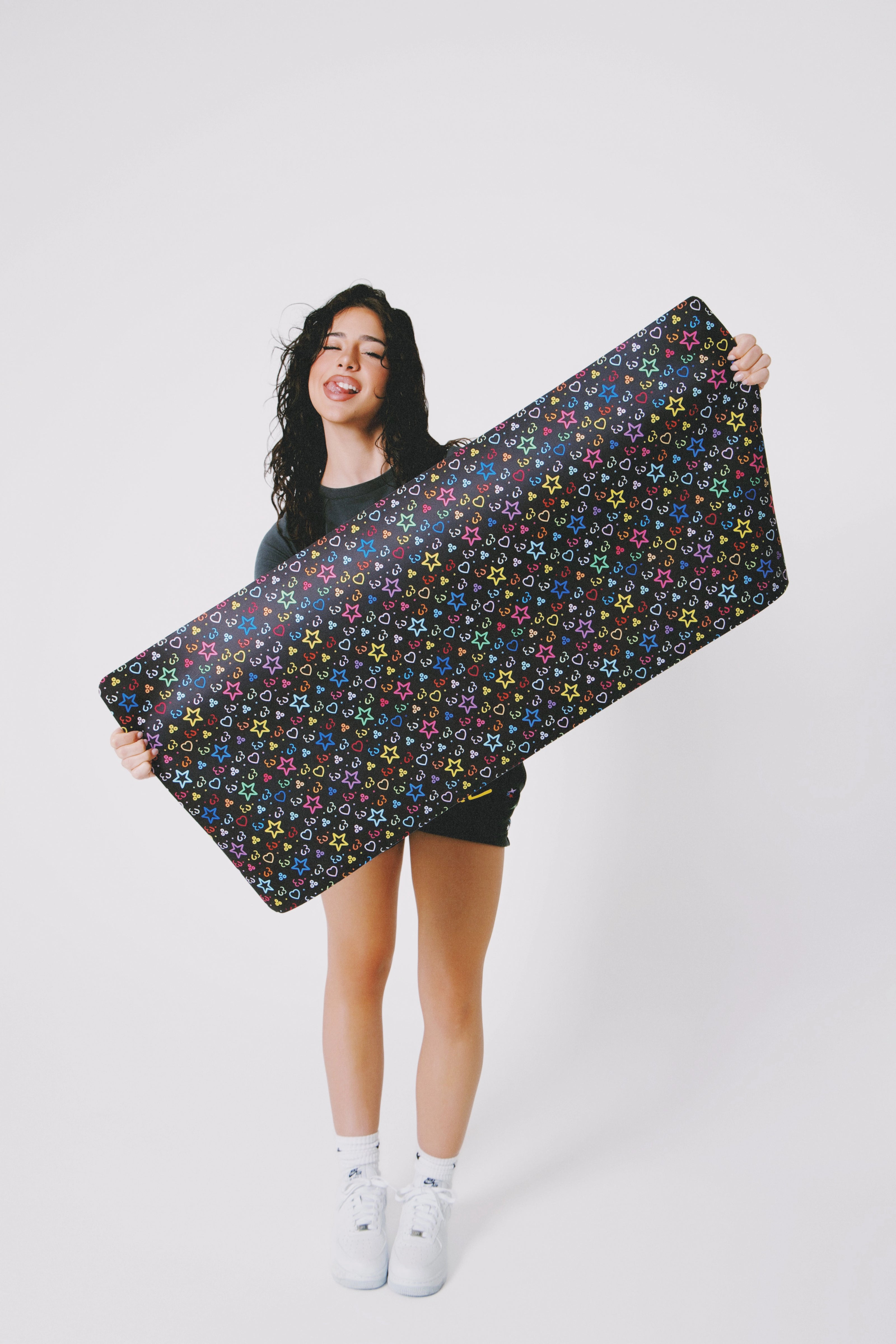 Model smiling and holding a large black mat with colorful star patterns, wearing a black crop top and shorts