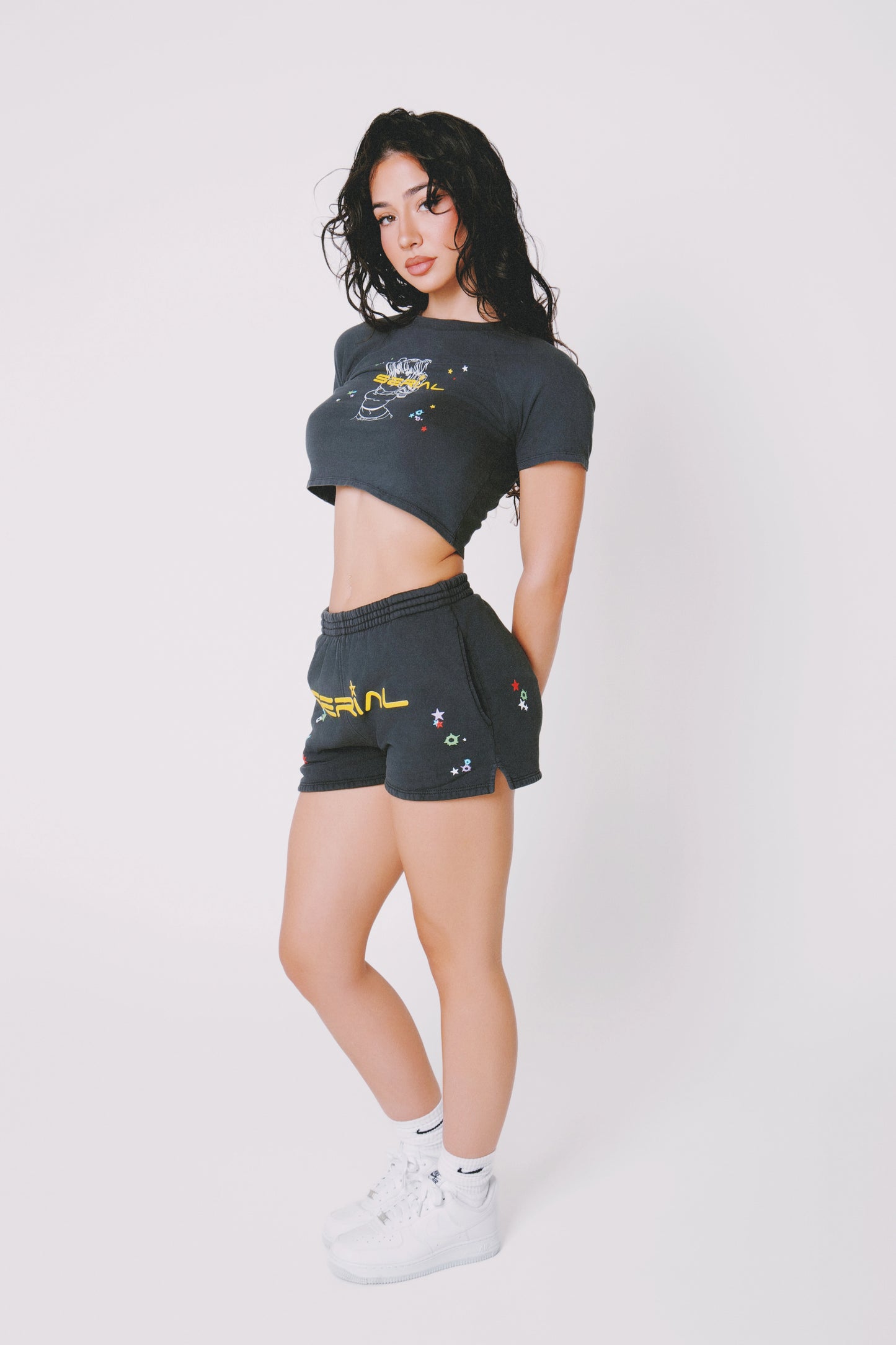 Model standing in profile wearing black 'SERIAL' branded shorts and crop top with colorful star designs, on a white background